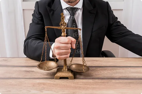 Man in black suit holding brass balance scale on wooden table in office.