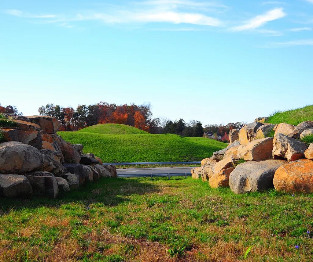 View of a grassy field framed by large rocks with a green hill and clear sky in the background.