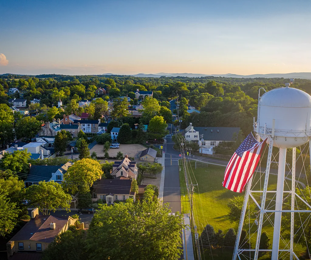 Aerial view of a small town with green trees, houses, and a water tower displaying an American flag.