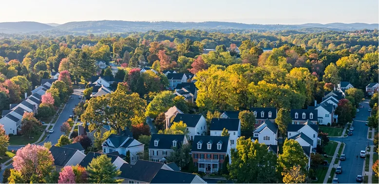 Aerial view of a suburban neighborhood with houses surrounded by autumn-colored trees and distant hills.