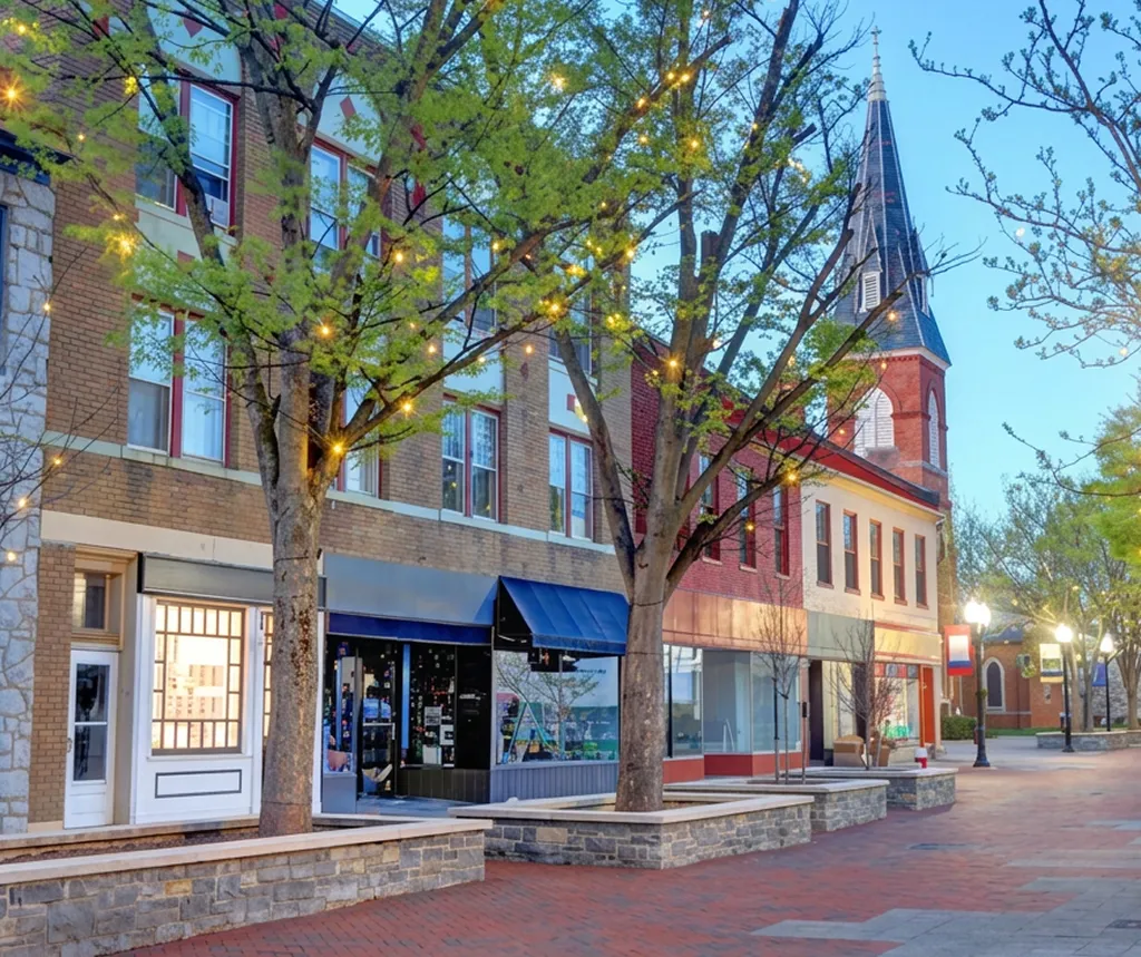 Downtown street scene with brick buildings, lit trees, and a church steeple at dusk.