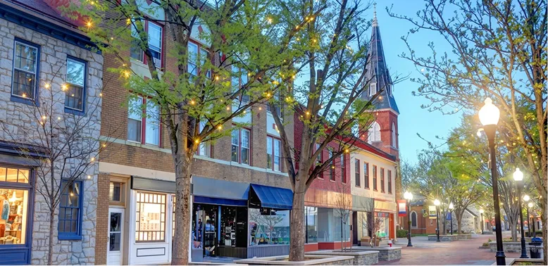 Downtown street scene at dusk with brick buildings, trees, and street lamps lit.