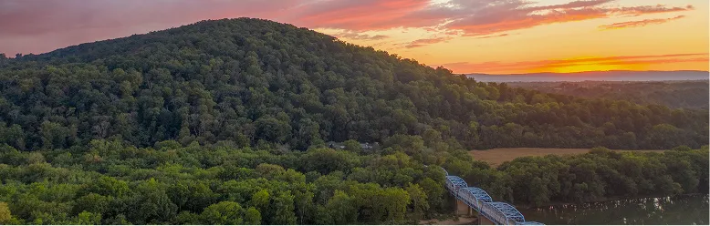 Panoramic view of a forested hill at sunset with a blue metal bridge crossing a river.
