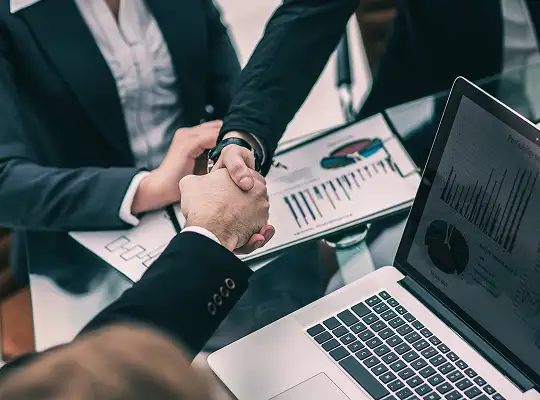 Two business professionals shaking hands over a table with charts and a laptop displaying financial graphs.