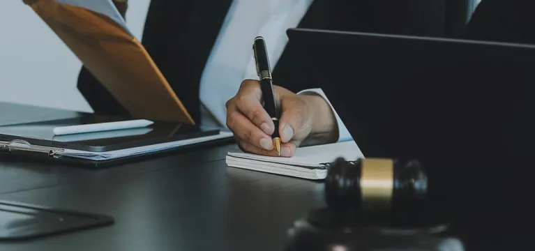 Professional in dark suit writing notes at desk with gavel and laptop nearby.
