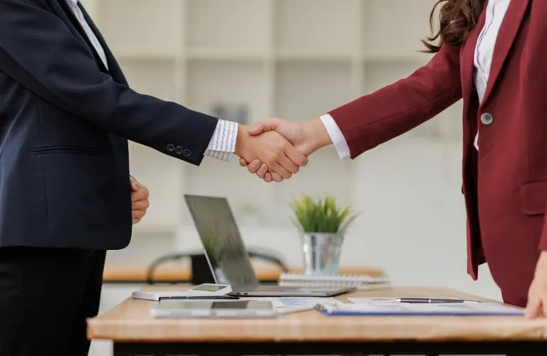 Two business professionals in formal suits shaking hands over a wooden office desk.
