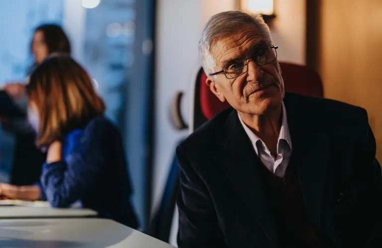 Elderly man with glasses wearing a dark suit seated indoors, two blurred women in background.