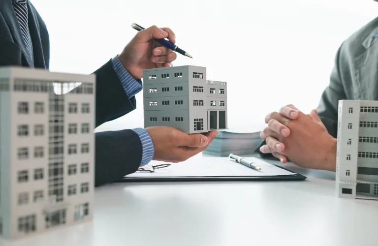 Two professionals in suits discussing a gray building model over a white table with documents.