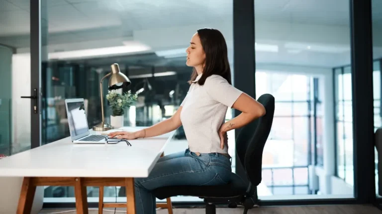 Woman in white shirt and jeans sitting at a desk, holding her lower back in discomfort.