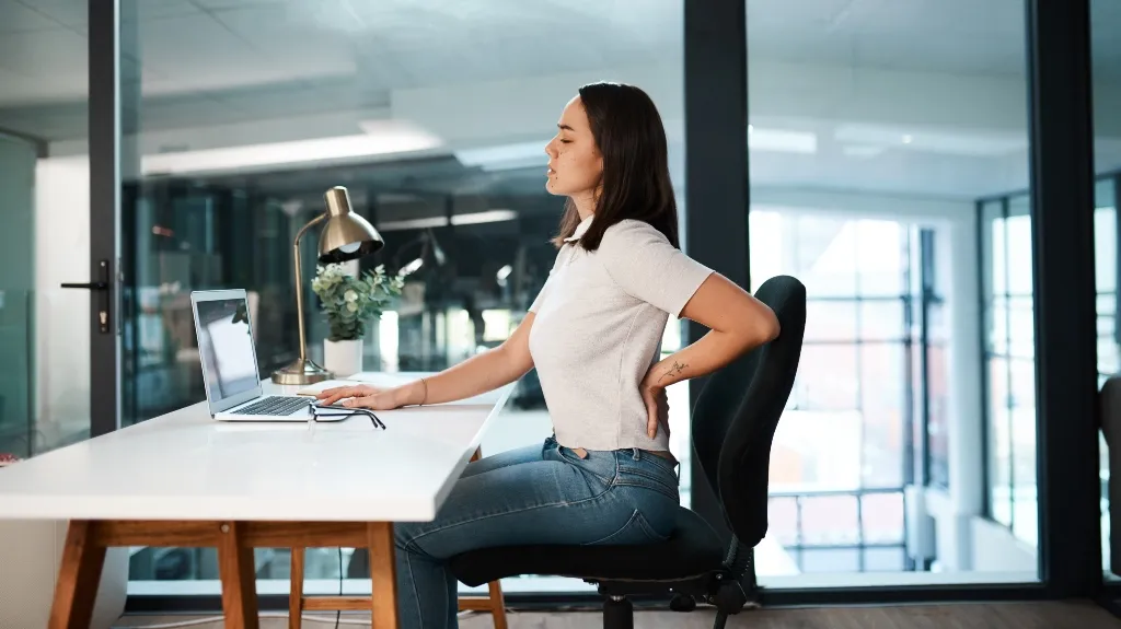 Woman in white shirt and jeans sitting at a desk, holding her lower back in discomfort.