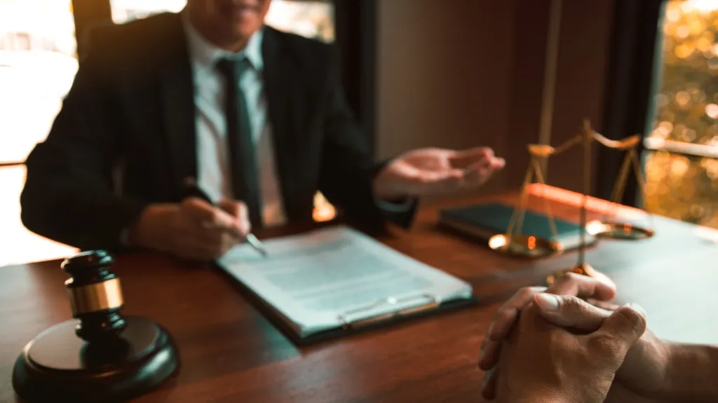 Two individuals in a legal consultation, with a gavel, scales, and documents on a wooden desk.