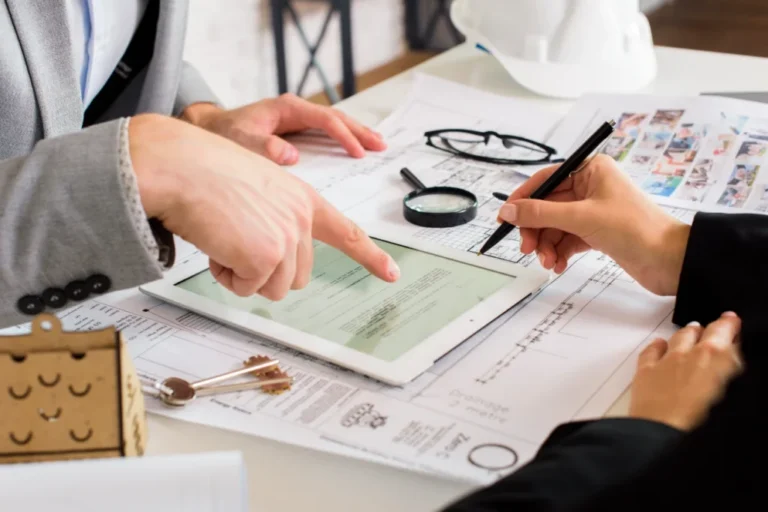 Two professionals reviewing documents and a tablet on a desk with architectural plans and a hard hat.