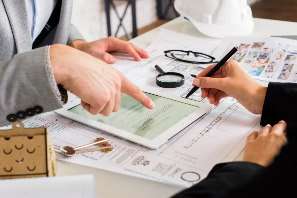 Two professionals reviewing documents and a tablet on a desk with architectural plans and a hard hat.